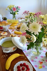 Brunch table with a white tablecloth with pink polka dots and on it flowers, pie, fruit, herbal teas, chocolate, meatballs, rice cakes and other delicious dishes