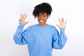 Young beautiful African American woman wearing blue knitted sweater against white wall showing and pointing up with fingers number nine while smiling confident and happy.