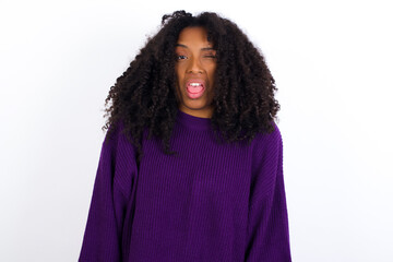 Young beautiful African American woman wearing knitted sweater against white wall,  sticking tongue out happy with funny expression. Emotion concept.