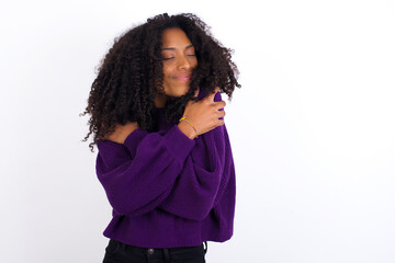 Young beautiful African American woman wearing knitted sweater against white wall,  Hugging oneself happy and positive, smiling confident. Self love and self care