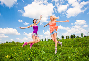 Two happy beautiful girls blonde and brunette twenty years old in colored dresses are jumping on a green field 