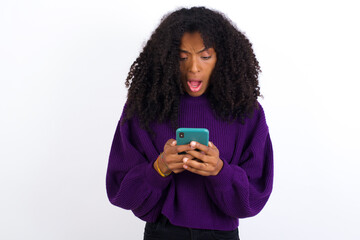 Focused Young beautiful African American woman wearing knitted sweater against white wall use smartphone reading social media news, or important e-mail