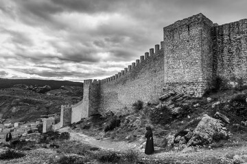 Monk walking in outdoor path, black and white scene with medieval old town wall.


