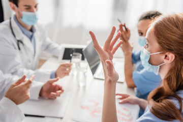 Redhead nurse in medical mask gesturing while talking to multiethnic colleagues at workplace on blurred background