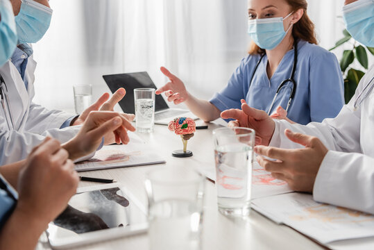 Doctors and nurses in medical masks talking while sitting at workplace with devices, pictures and glasses of water on blurred foreground
