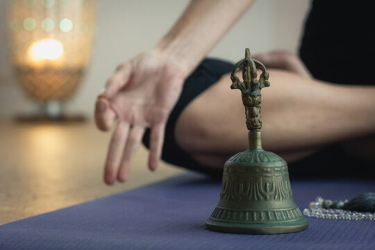 Detail of metal bell on yoga mat and body of woman sitting in padmasana with lamp on background. Yogi in lotus pose doing meditation ritual ceremony in studio. Zen, spiritual concepts