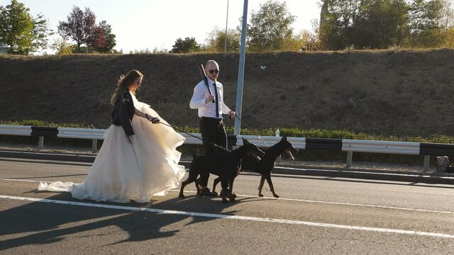 The bride and groom are walking along the highway holding two Dobermans on leashes. The groom is holding a baseball bat.