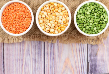 Top view of three mugs filled to the brim with yellow, green peas and lentils. The mugs stand on a matting that lies on a wooden table