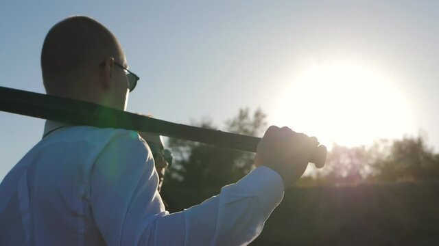 A couple of newlyweds on the highway are in the sunglasses and looking into the distance. The groom leans a baseball bat on his shoulder.