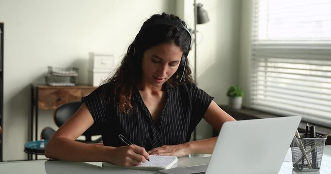 Woman sit at desk wear headset e-learning using laptop and videocall app, listens tutor, writes information on copybook, gain new knowledge remotely. Video conference event distance education concept