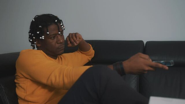 Relaxed young black man watching tv at home, changing the channels using remote controller. High quality 4k footage