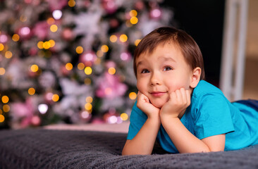 Fototapeta na wymiar Toddler boy in blue shirt lying on belly in room and dreaming. Blurred decorated Christmass tree in background. Christmas miracle, dreams, family winter holidays and New Year resolutions concept