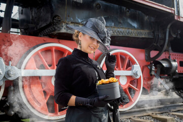 Beautiful girl in a black dress and hat near an old steam locomotive and big iron wheels. Blond beauty. Vintage portrait of the last century, retro journey.