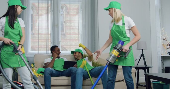 Beautiful positive hardworking two female cleaner are dissatisfied that their male cheerful multiracial colleagues don't help them but watching something on tablet device and having fun together