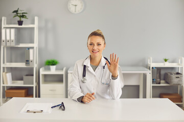 Young smiling blonde woman doctor nurse therapist sitting and greeting patient with hand online