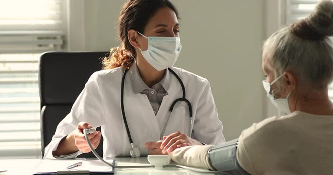 Nurse female doctor use tonometer checking up measuring pressure of retired elderly female patient during visit at hospital, people wear face mask due corona virus pandemic outbreak, eldercare concept