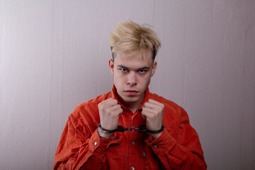 A handcuffed teenager with an angry expression stretches out his hands in front of a gray...