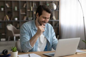 Overjoyed young Caucasian man look at laptop screen talk on video call on gadget. Smiling millennial male sit at desk at home office work distant on computer, have webcam online communication.
