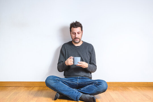 caucasian man on his 40s dressing casual and sitting on the wooden floor of an empty apartment with a blue coffee mug on his hands
