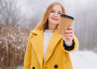 Happy young Woman holds paper coffee cup over nature background, trendy color of year 2021 - illuminatiing yellow. Take away or delivery concept. Copy space. Place for your text or logo on mug