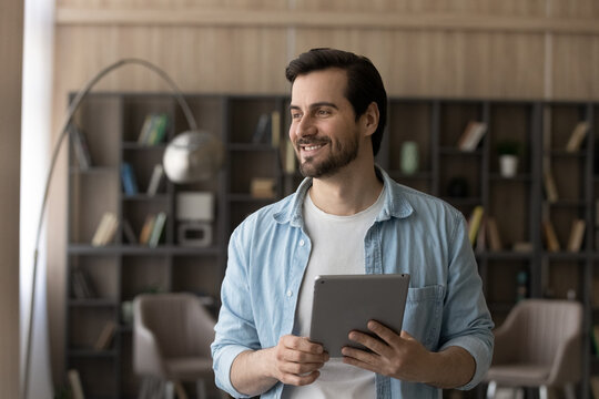 Happy millennial Caucasian man look in distance using modern tablet at home. Smiling young male user or client hold pad device, thinking or pondering. Online distant communication, technology concept.