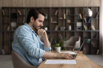 Pensive serious millennial Caucasian man sit at home office desk work on laptop online....