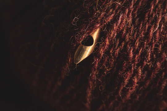 Macro photography of a golden 5 millimeter punch needle tip coming out from a dark red wool ball.