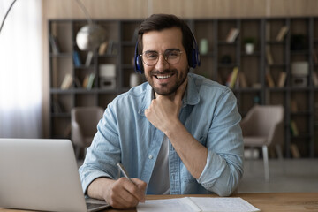 Portrait of smiling millennial male student in headphones sit at desk at home study online on...