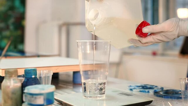 Pouring epoxy resin into a plastic cup from a canister. Preparation of material for creation in an art workshop. Close-up.
