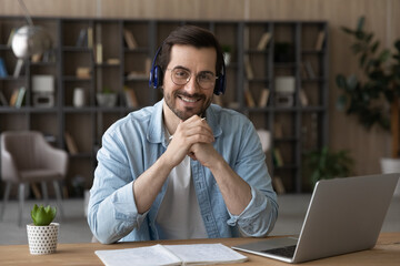 Portrait of smiling young Caucasian man in earphones sit at desk study online on laptop. Happy...