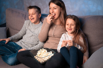 mom and kids sit on the couch in the evening, eat popcorn, watch TV and laugh.