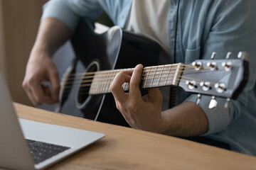 Fototapeta na wymiar Crop close up of male music coach or tutor play guitar have online video lesson on laptop at home studio. Man artist or singer use musical instrument record new song or single. Hobby concept.