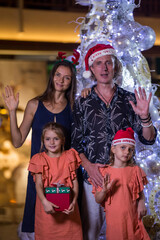 Happy family with two daughters standing by Christmas tree holding gift box present, raising hands and waving. Merry Christmas and happy holidays