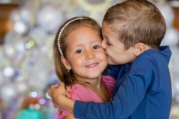Cute boy standing by Christmas tree, hugging and kissing little sister. Merry Christmas and happy holidays