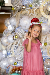 Portrait of happy cute little girl wearing Santa hat standing by Christmas tree, waving and greeting with her hand. Merry Christmas and happy holidays