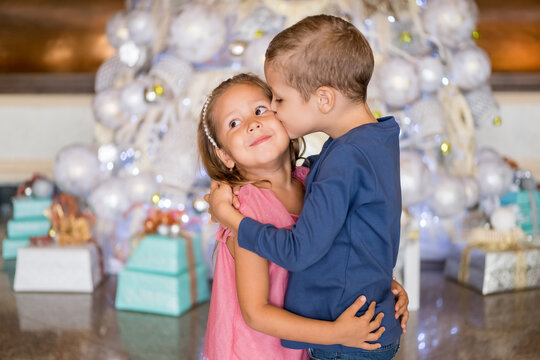 Cute boy standing by Christmas tree, hugging and kissing little sister. Merry Christmas and happy holidays