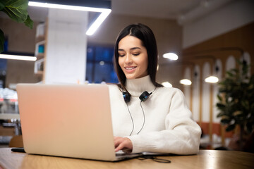 A young smiling businesswoman with headphones works on a laptop in a co-working space, a young manager in a headset consults a client online or listens to music using a computer in a shared office