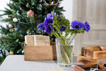 A bouquet of blue anemones in a glass vase on a table covered with a gray tablecloth in front of a Christmas tree as a room decoration