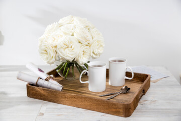 Bouquet of white roses in a round glass vase on a tray with two cups of tea and spoons in the interior