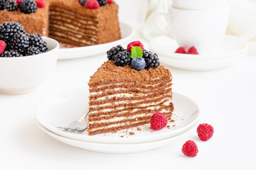 Chocolate honey cake with cream and fresh berries on top on a white plate on a light concrete background. Copy space.