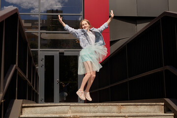 A happy young girl in a skirt jumps up. Black background with a red stripe. A large building and the sky and clouds reflected in it.