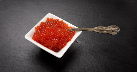 fresh grained red chum salmon caviar in a white ceramic bowl on a black slate stone background