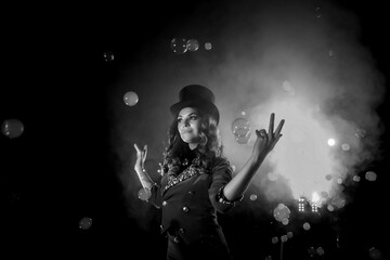 Female magician makes show with soap bubbles, an illusionist in theatrical clothes, on black background. Woman actress in stage costume and top hat on her head. Concept of performance. Copy space