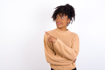 Obraz na płótnie Canvas Portrait of Young beautiful African American woman wearing knitted sweater against white wall, posing on camera with tricky look, presenting product with index finger. Advertisement concept.