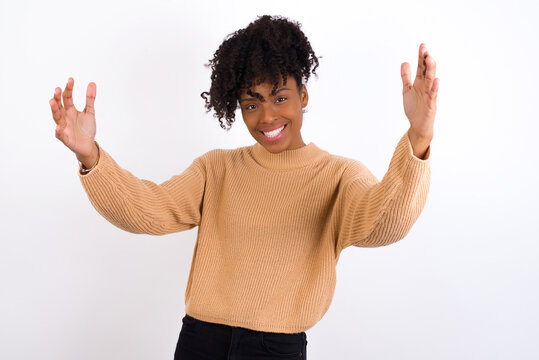 Cheerful Young beautiful African American woman wearing knitted sweater against white wall, making a welcome gesture raising arms over head.