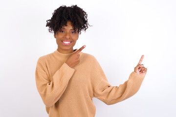 Positive Young beautiful African American woman wearing knitted sweater against white wall with beaming smile pointing with two fingers and looking on empty copy space. Advertisement concept.