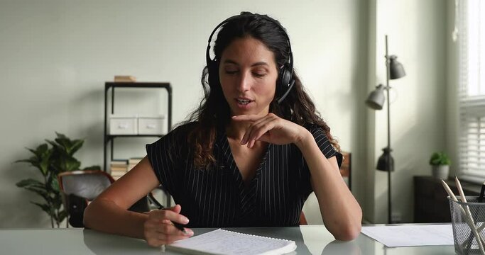 Woman in headset hold pen ready make notes and receive new knowledge, sit at table looks at camera start conversation talk use device and video call app. E-learn, webinar, distant consultation concept