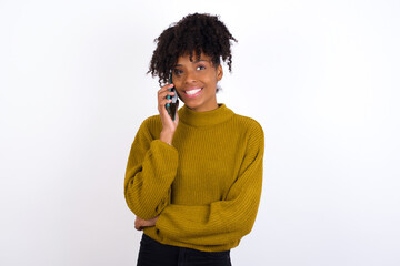 Portrait of a smiling Young beautiful African American woman wearing knitted sweater against white wall talking on mobile phone. Business, confidence and communication concept.
