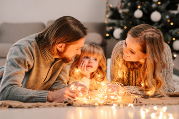 Happy family with daughter lies on the floor and cozy talks by the light of garlands against the background of Christmas tree