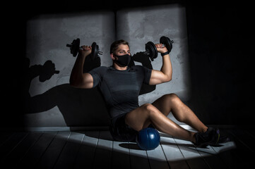 Obraz na płótnie Canvas Male athlete wearing protective face mask and training with dumbbell in gym. Workout in gym after pandemic